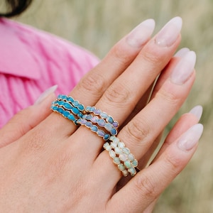 May include: Three gold-toned rings with colorful, oval-shaped stones are displayed on a hand. The rings feature blue, purple, and white stones. The rings are stacked, creating a layered effect. The background is blurred, with a pink sweater visible.