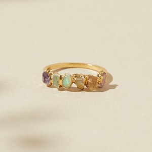 May include: A gold ring adorned with a series of colourful, irregularly shaped gemstones. The gemstones display hues of purple, green, and yellow. The ring is set against a soft, neutral backdrop, accentuating the vibrant colours of the stones.