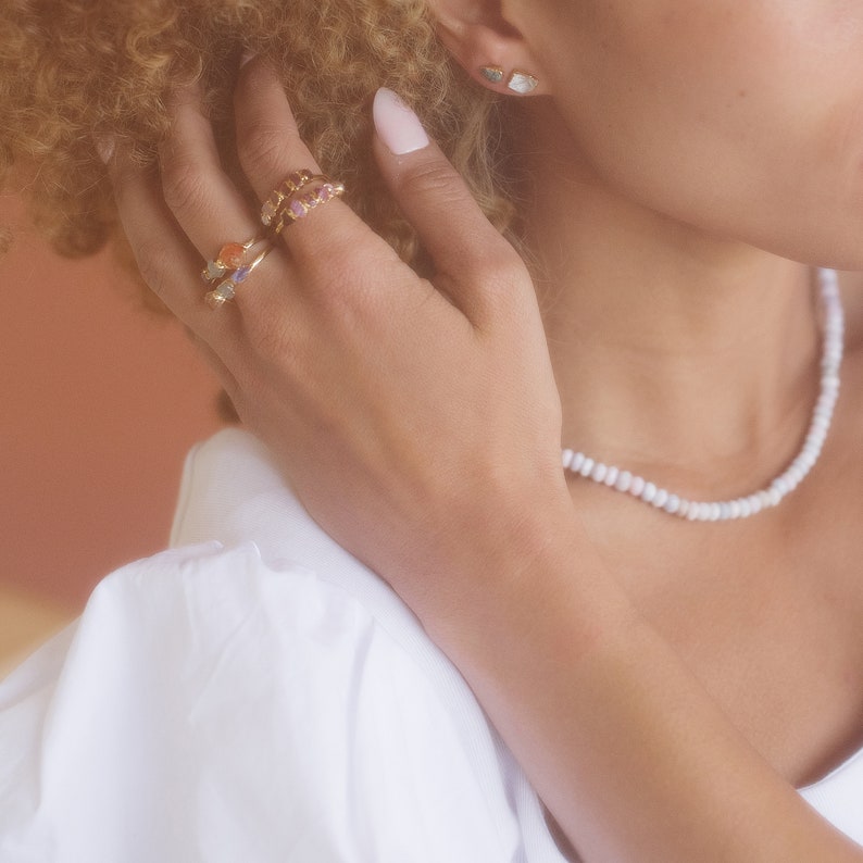 Pu&ograve; includere: Due anelli in oro con piccole pietre preziose multicolori indossati sulle dita, un paio di piccoli orecchini a lobo con pietre di colore chiaro e una semplice collana di perline bianche indossata al collo, presentati come accessori di gioielleria.