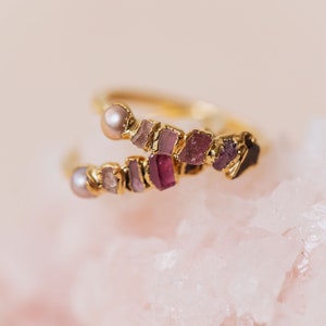 May include: A gold bypass ring featuring two rows of raw, multi-tonal pink and purple gemstones interspersed with small, round, lustrous pearls. The stones vary in shade from pale blush to deep magenta, set along a polished gold-toned metal band.