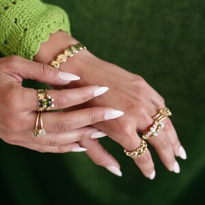 Puede incluir: Primer plano de manos con anillos de oro con varias piedras preciosas. Los anillos est&aacute;n apilados en varios dedos. Las u&ntilde;as est&aacute;n pintadas de blanco y son largas y de forma almendrada.