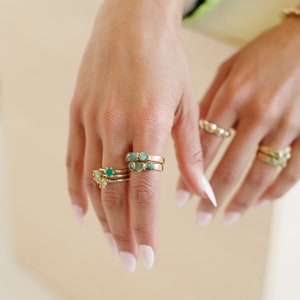 May include: A hand wearing multiple gold rings with green gemstones. The rings are stacked on the middle finger.