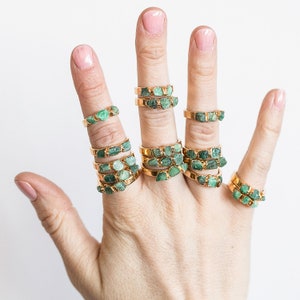 May include: A hand wearing multiple gold rings with green emerald stones. The rings are stacked on each finger.