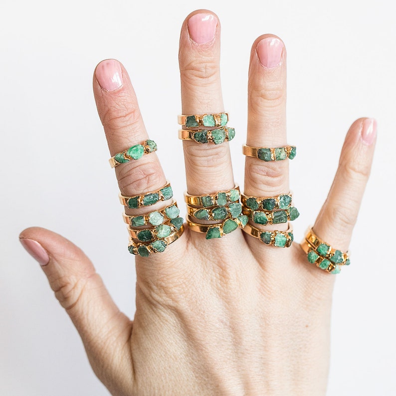 May include: A hand wearing multiple gold rings with green emerald stones. The rings are stacked on each finger.