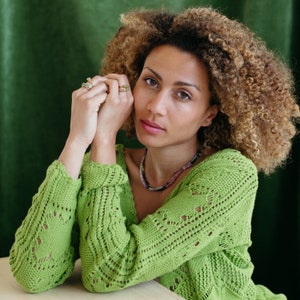 May include: A woman with curly brown hair wearing a green crocheted cardigan with a delicate pattern. She is wearing several rings on her fingers and a necklace with small stones.