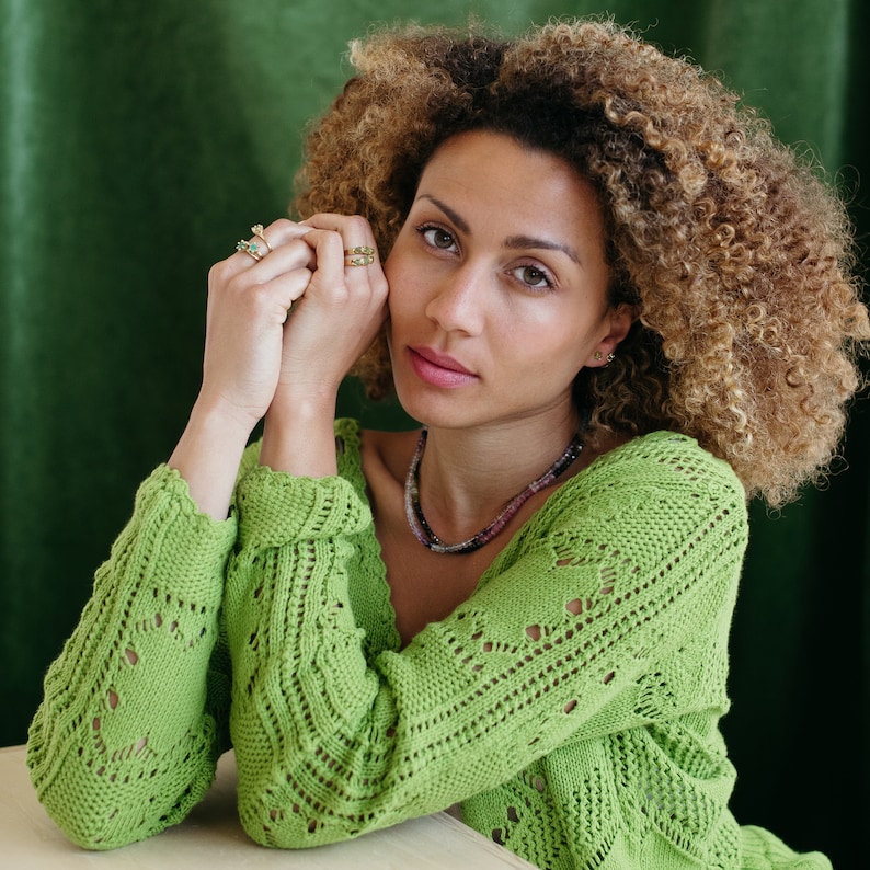May include: A woman with curly brown hair wearing a green crocheted cardigan with a delicate pattern. She is wearing several rings on her fingers and a necklace with small stones.