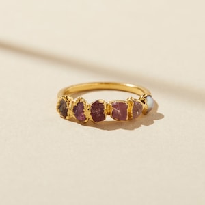 Pu&ograve; includere: Un anello in oro con una fila di sei pietre preziose grezze e non tagliate in varie tonalit&agrave; di rosa, viola e rosso scuro, incastonate in una fascia d'oro testurizzata. Le pietre presentano bordi irregolari e frastagliati.