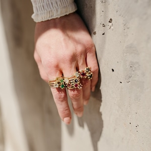 Puede incluir: Una mano con una pila de anillos de oro con piedras preciosas de colores. Los anillos presentan una variedad de piedras, incluyendo esmeralda, rub&iacute;, zafiro y diamante.