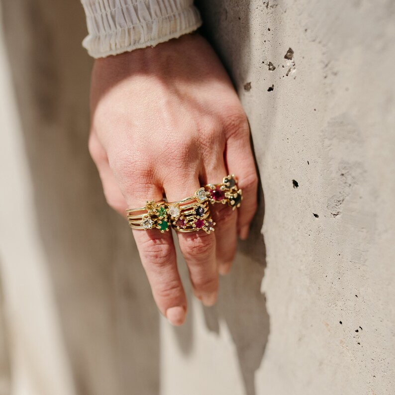 Puede incluir: Una mano con una pila de anillos de oro con piedras preciosas de colores. Los anillos presentan una variedad de piedras, incluyendo esmeralda, rub&iacute;, zafiro y diamante.