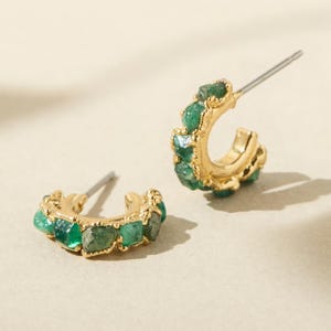 May include: A pair of gold-colored hoop earrings with a row of rough-cut, emerald-green gemstones. The earrings have a post closure and are set against a neutral background.