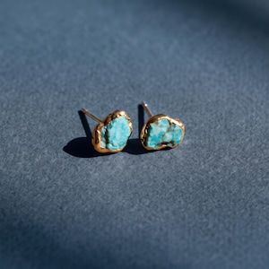 Könnte beinhalten: Ein Paar Ohrstecker mit rohen, unregelmäßigen Türkissteinen, die natürliche blaue und braune Maserungen aufweisen. Jeder Stein ist in eine polierte, goldfarbene Metallfassung mit einem Stift gefasst und zeigt eine raue, organische Struktur.