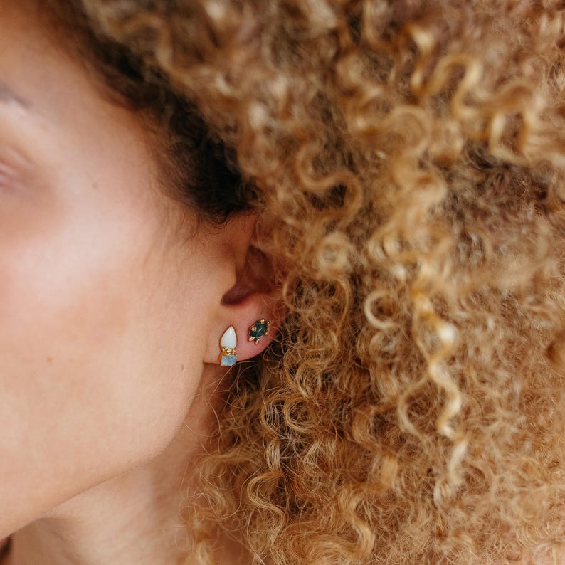 Close-up of opal aquamarine earrings on a model with curls. Rough gemstone studs in a gold setting.