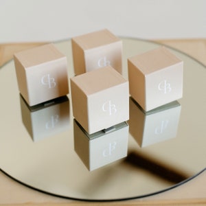 May include: Four beige boxes with the letters "CB" printed in white on the top of each box. The boxes are arranged in a square on a mirrored surface.