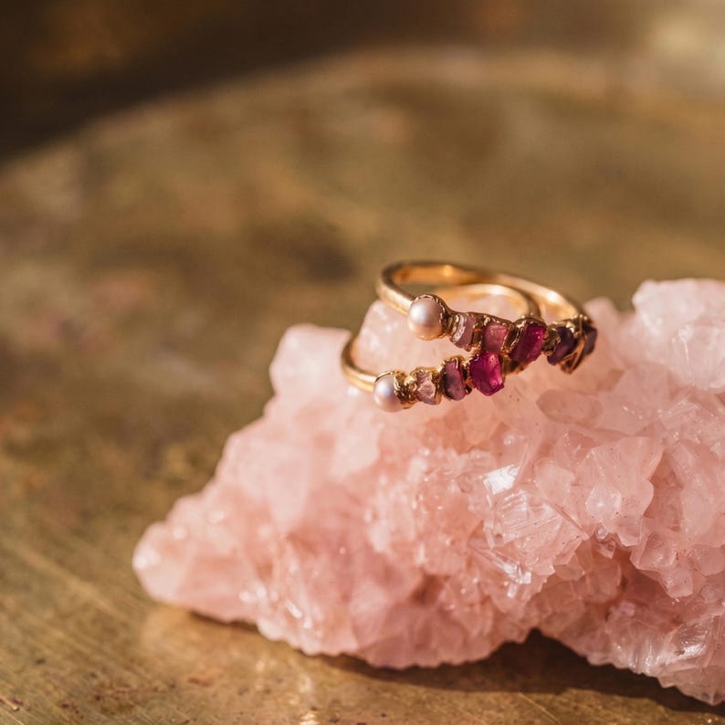 Pu&ograve; includere: Un anello color oro con una piccola perla bianca e una serie di pietre preziose grezze color rosa scuro e magenta disposte lungo la fascia. L'anello poggia su un grande pezzo frastagliato di cristallo di quarzo rosa pallido e traslucido.