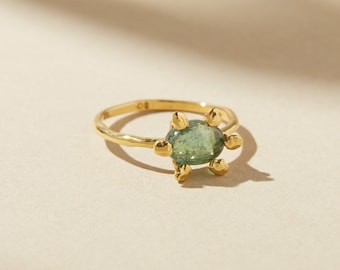 May include: A gold ring with a central oval-shaped, pale green gemstone. The ring has six small, gold-coloured accents around the gemstone, resembling a turtle. The band has a hammered texture. The ring is a piece of jewellery.