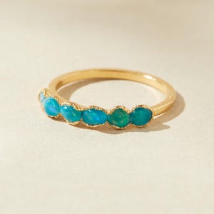 Peut inclure: Une bague en or ornée d'une rangée de pierres précieuses ovales bleu-vert. Le corps de la bague est en or fin et poli. Les pierres sont serties côte à côte, créant une ligne continue de couleur. Les pierres ont une légère irisation.