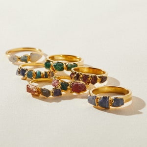May include: A set of six gold rings with different colored gemstones. Each ring features a cluster of small, irregular shaped stones. The stones are a mix of green, blue, purple, and pink.