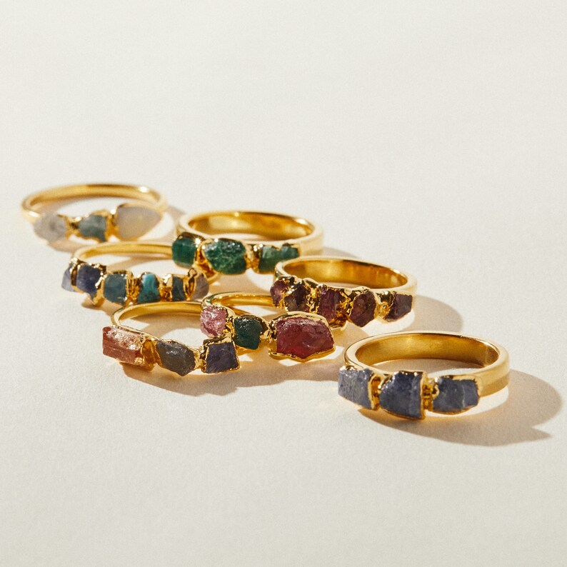 May include: A set of six gold rings with different colored gemstones. Each ring features a cluster of small, irregular shaped stones. The stones are a mix of green, blue, purple, and pink.