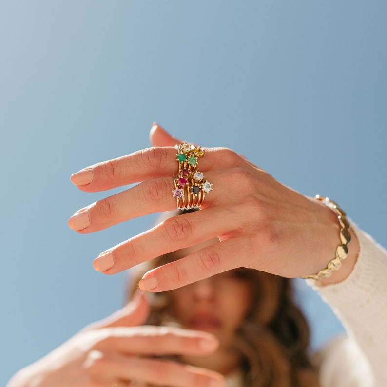 Puede incluir: Una mano lleva una pila de anillos de oro con piedras preciosas de colores. Los anillos est&aacute;n apilados en el dedo medio. La mano est&aacute; llegando hacia la c&aacute;mara.