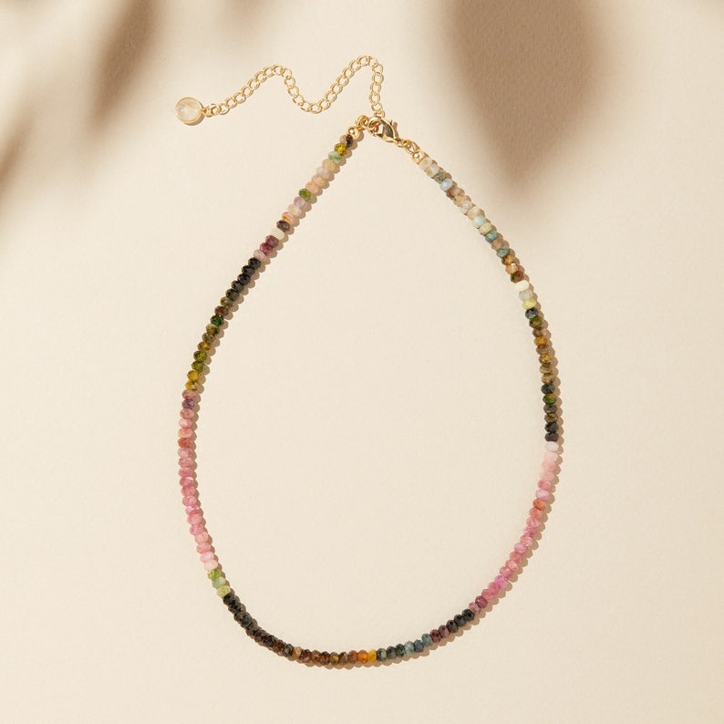 May include: A delicate gold chain necklace with a variety of small, colorful gemstones. The necklace features a mix of pink, green, and black gemstones.