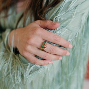 Pu&ograve; includere: Un anello in oro con una fila di tre pietre preziose verdi rettangolari e una fila di tre pietre rosa di forma ovale disposte verticalmente. La fascia &egrave; sottile e metallica, mostrando una combinazione di tonalit&agrave; verdi e rosa.