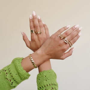 Puede incluir: Un par de manos que llevan m&uacute;ltiples anillos de oro con varias piedras preciosas, incluida una piedra verde, en dos dedos de cada mano. Las manos llevan un brazalete de oro con una cadena de peque&ntilde;os eslabones redondos en una mu&ntilde;eca. Las manos est&aacute;n sobre un fondo beige claro.