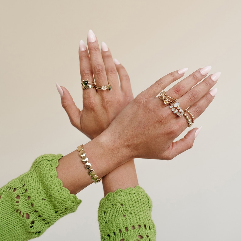 Puede incluir: Un par de manos que llevan m&uacute;ltiples anillos de oro con varias piedras preciosas, incluida una piedra verde, en dos dedos de cada mano. Las manos llevan un brazalete de oro con una cadena de peque&ntilde;os eslabones redondos en una mu&ntilde;eca. Las manos est&aacute;n sobre un fondo beige claro.