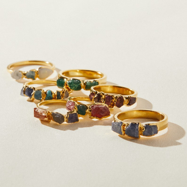 May include: A set of six gold rings with different colored gemstones. Each ring features a cluster of small, irregular shaped stones. The stones are a mix of green, blue, purple, and pink.