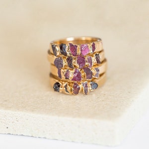 Könnte beinhalten: Ein Stapel goldener Ringe mit rosafarbenen und violetten Edelstein-Akzenten. Jeder Ring hat eine einzigartige Anordnung von rohen Steinen.