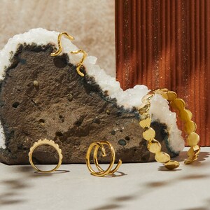 Puede incluir: Cuatro anillos de oro sobre una superficie texturizada. Dos anillos son bandas simples, uno es un dise&ntilde;o de doble bucle y otro es una banda gruesa con un borde festoneado.