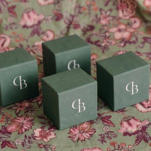 Pu&ograve; includere: Quattro scatole regalo quadrate color verde salvia opaco disposte in gruppo. Ogni scatola presenta sulla parte anteriore un logo monogramma bianco stilizzato composto dalle lettere C e B intrecciate. Le scatole hanno una finitura esterna liscia.