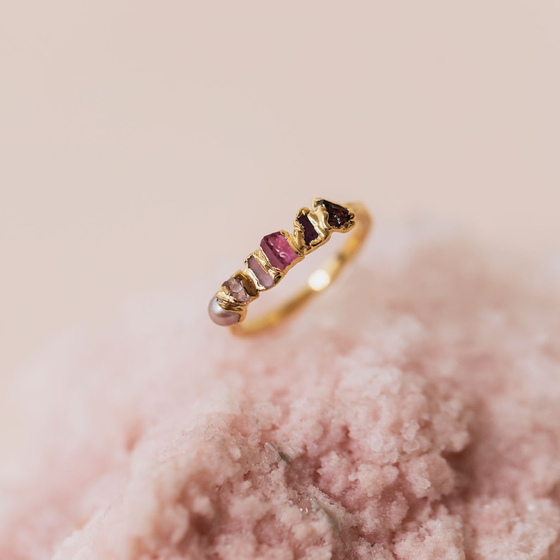 Pu&ograve; includere: Un anello in oro con una fila di piccole pietre preziose grezze multicolori e una singola perla rotonda. Le pietre sfumano dal rosa chiaro e malva al magenta intenso e rosso scuro, incastonate in una fascia d'oro dalla superficie ruvida.