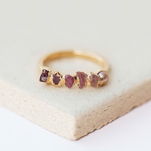 Pu&ograve; includere: Un anello in oro con una fila di cinque pietre preziose grezze nelle tonalit&agrave; del rosso scuro, magenta e rosa, accentuato da una singola piccola perla rotonda rosa pallido all'estremit&agrave; della montatura. L'anello &egrave; in oro liscio e lucido.