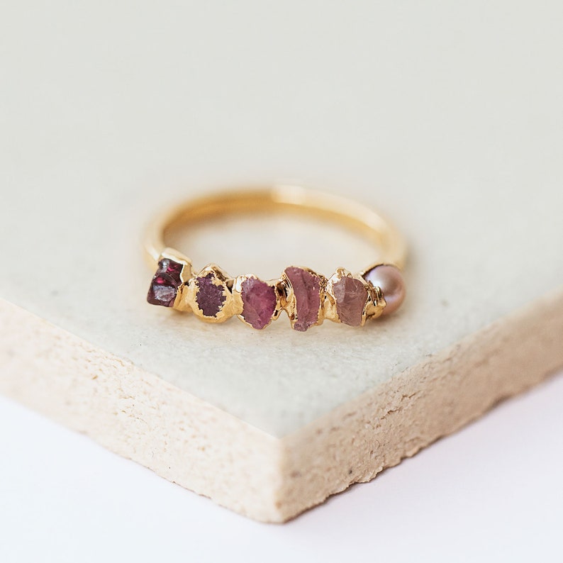 Pu&ograve; includere: Un anello in oro con una fila di cinque pietre preziose grezze nelle tonalit&agrave; del rosso scuro, magenta e rosa, accentuato da una singola piccola perla rotonda rosa pallido all'estremit&agrave; della montatura. L'anello &egrave; in oro liscio e lucido.