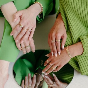 Puede incluir: Dos manos con m&uacute;ltiples anillos de oro con piedras preciosas de colores. Los anillos est&aacute;n apilados en cada dedo y presentan una variedad de estilos, incluyendo bandas, anillos delicados y piezas llamativas.