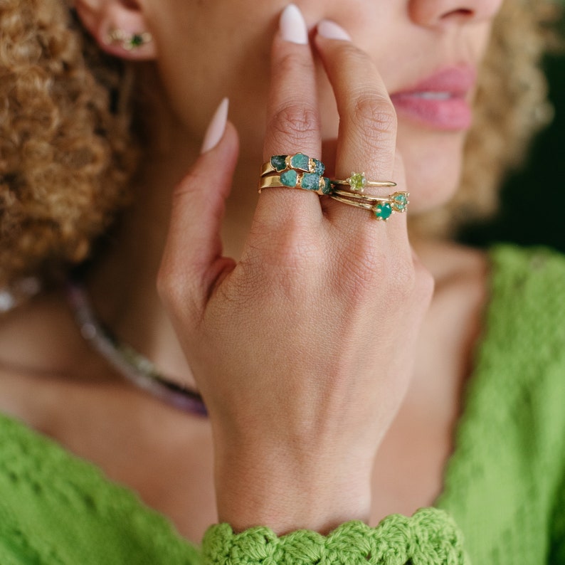 May include: A hand wearing four gold rings with green gemstones. The rings are stacked on the ring finger. The hand is wearing a green knitted sweater.