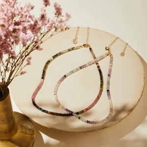 May include: Two gold necklaces with colorful gemstone beads. The necklaces are layered on a white surface.