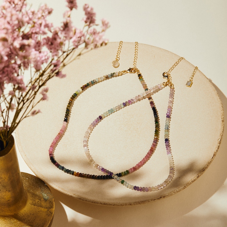 May include: Two gold necklaces with colorful gemstone beads. The necklaces are layered on a white surface.