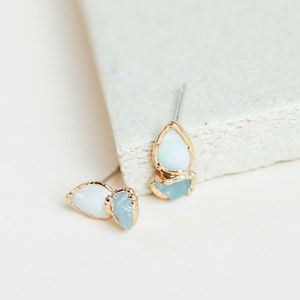 Close-up of rough opal and aquamarine studs on a white stone block. Dainty gold earrings with raw crystals.