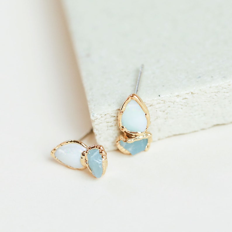 Close-up of rough opal and aquamarine studs on a white stone block. Dainty gold earrings with raw crystals.