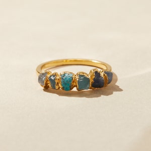 May include: Gold ring with five different colored gemstones set in a row. The stones are a mix of blue, green, and gray.