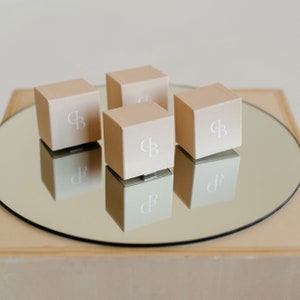 Puede incluir: Cuatro peque&ntilde;as cajas cuadradas de color beige con un logotipo "CB" blanco en cada una. Las cajas est&aacute;n dispuestas en c&iacute;rculo sobre una superficie de espejo.