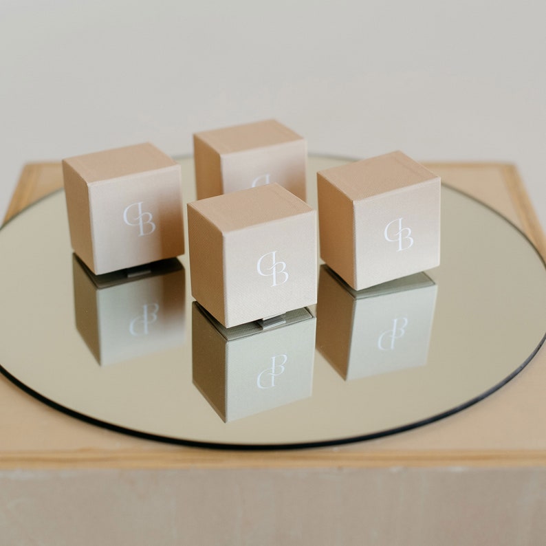 Puede incluir: Cuatro peque&ntilde;as cajas cuadradas de color beige con un logotipo "CB" blanco en cada una. Las cajas est&aacute;n dispuestas en c&iacute;rculo sobre una superficie de espejo.