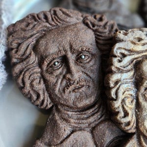 May include: Close-up of a detailed, brown sculpture of a man's face with curly hair and a moustache. The sculpture has a textured surface and is likely made of a dense material. The image suggests a bust or relief sculpture.