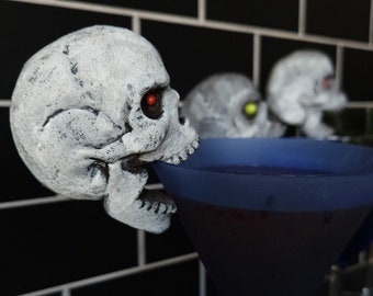 Puede incluir: Un cóctel de Halloween espeluznante. Una calavera blanca decora el borde de una copa de martini azul llena de un líquido morado oscuro. Dos calaveras más están en el fondo, con ojos rojos y amarillos. El fondo es una pared de azulejos negros.