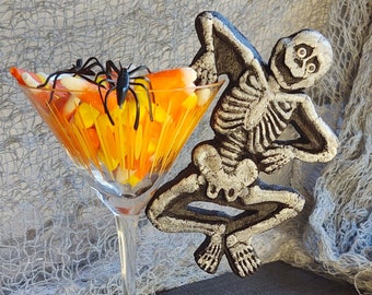 Puede incluir: Un vaso de martini de vidrio lleno de maíz dulce naranja y blanco, con una araña negra en el borde. Una figura de esqueleto se apoya en el vaso, con los brazos levantados.