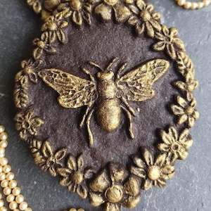 May include: Two dark brown round cookies decorated with gold and silver bees and gold flowers. The cookies are on a gray surface with a pearl necklace.