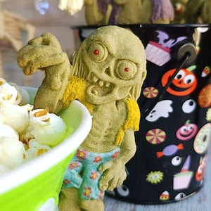 May include: Two green zombie cookie decorations with red eyes and a toothy grin. The zombie on the left is holding a green bucket filled with popcorn. The zombie on the right is holding a black bucket with a Halloween theme.