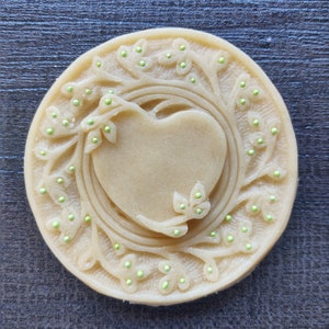 May include: A round, light brown cookie with a heart-shaped design in the center. The heart is surrounded by a decorative border of leaves and small green dots.