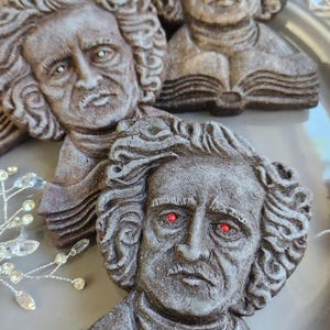 Puede incluir: Primer plano de varias galletas con forma de rostro de Edgar Allan Poe, esculpidas en marr&oacute;n oscuro, con ojos rojos, dispuestas sobre un plato gris. Las galletas est&aacute;n detalladas con pelo rizado y libros abiertos. Se ven algunos detalles decorativos de cristal.